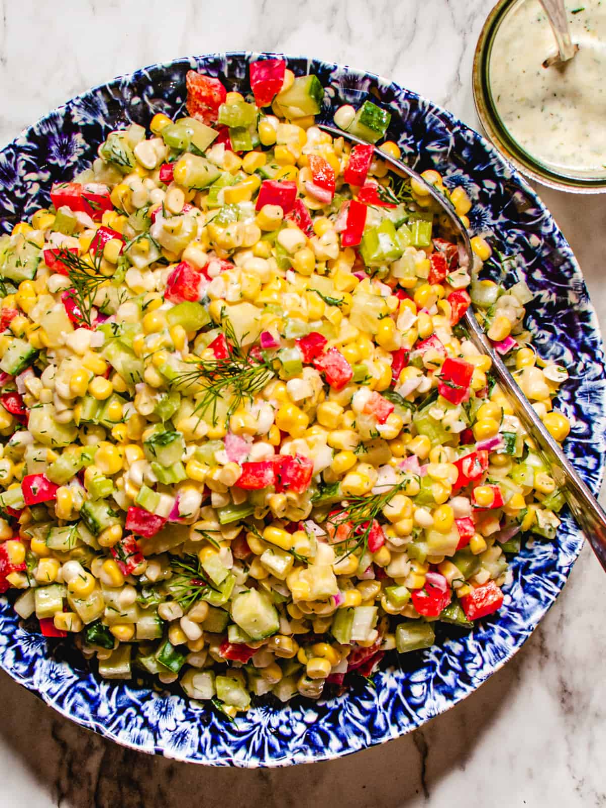 Corn salad in blue and white bowl on marble background with jar of creamy dill dressing.