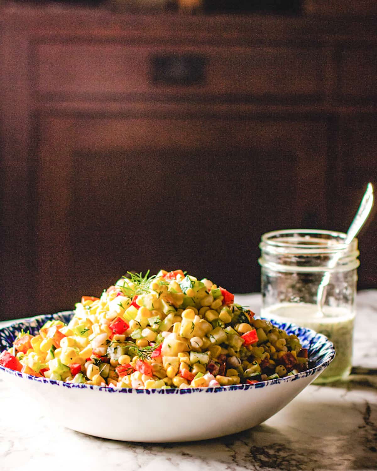 Corn salad in a blue and white bowl, black background and jar of creamy dill dressing.