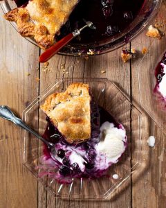 Slice of blueberry pie with scoop of ice cream on clear plate on wood. Overhead shot