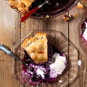Slice of blueberry pie with scoop of ice cream on clear plate on wood. Overhead shot