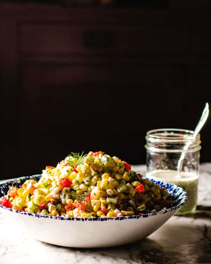 Corn salad in a blue and white bowl, black background and jar of creamy dill dressing