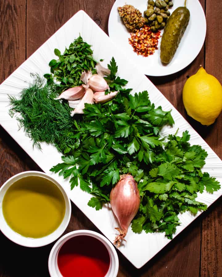 all ingredients for making salsa verde. fresh herbs, lemon, olive oil, mustard, capers, shallot, pickle and vinegar
