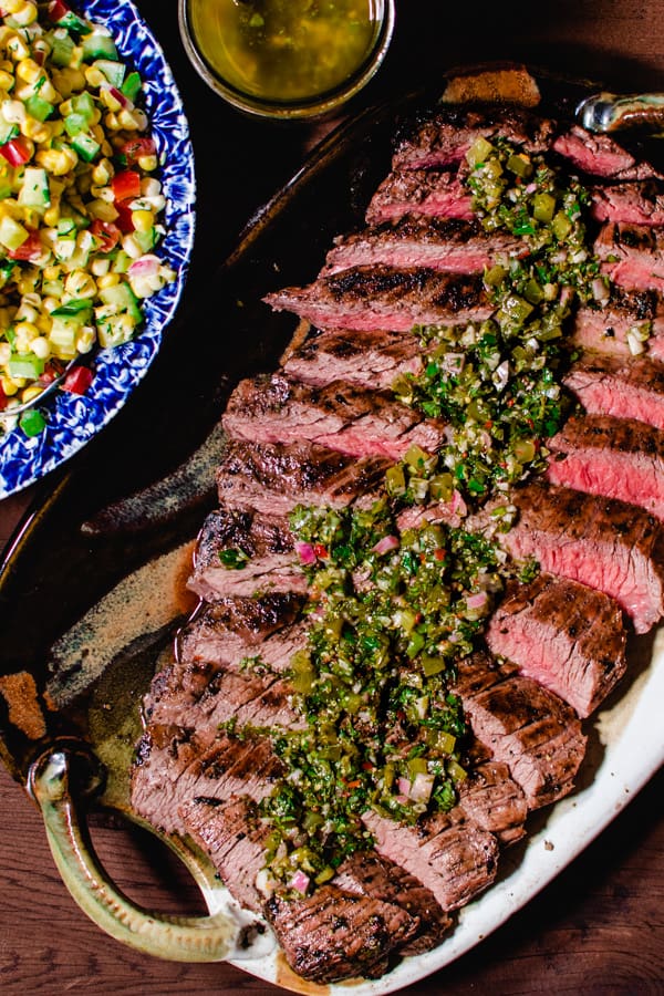 grilled flank steak with salsa verde on ceramic platter with corn salad and jar of salsa in the background