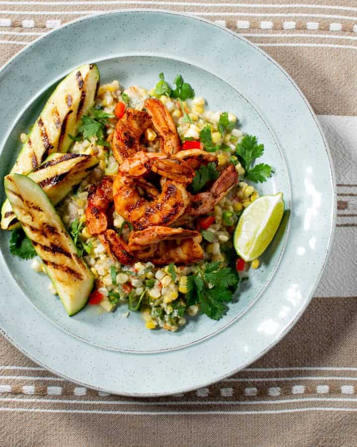 Grilled shrimp with coconut creamed corn on blue plate. Garnished with grilled zucchini and fresh lime