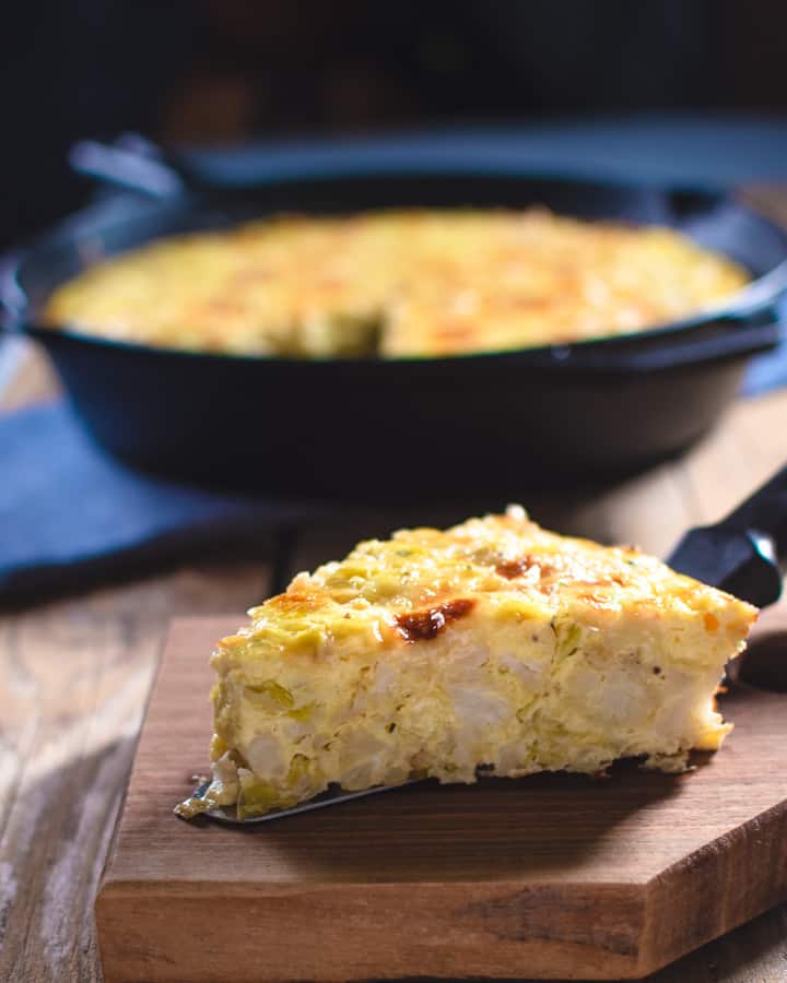 slice of cauliflower frittata on wood board with cast iron pan in background