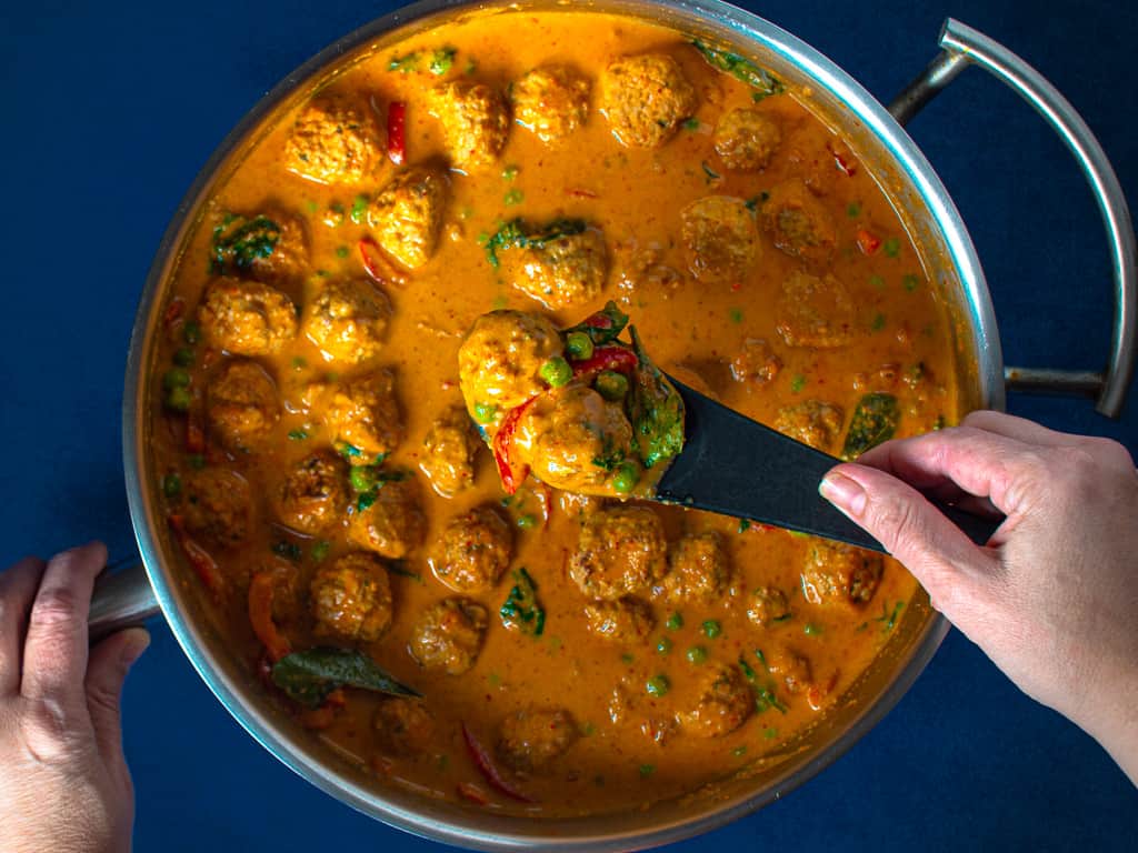 a pan of meatballs in red curry peanut sauce with a black spoon holding 3