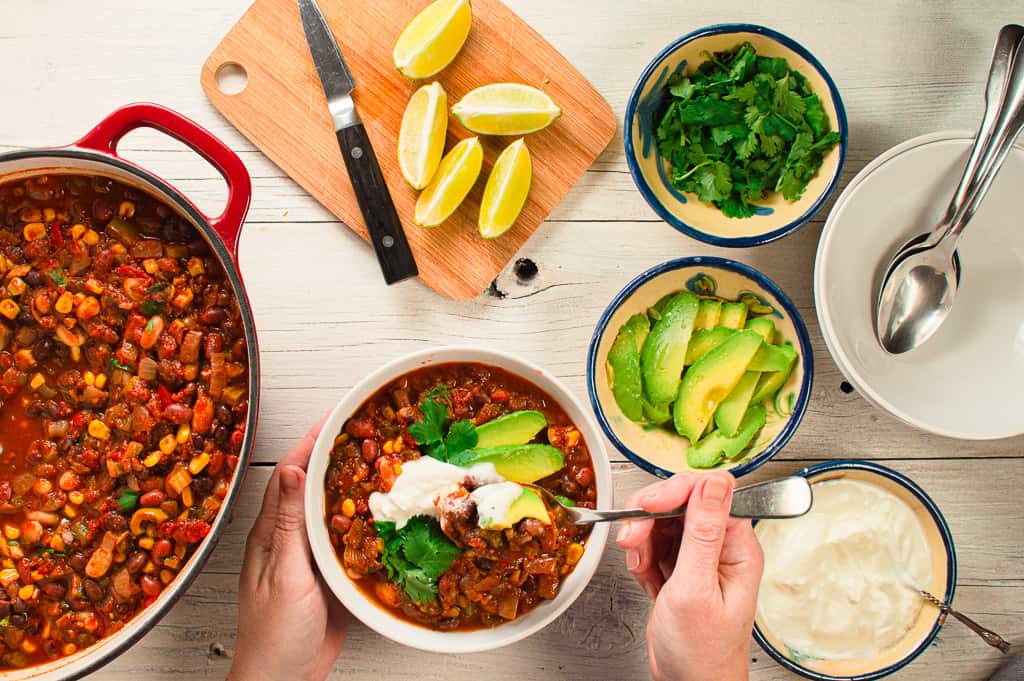 one bowl of vegetarian chili with the pot on the side. Small dishes of toppings. Avocado, cilantro and limes