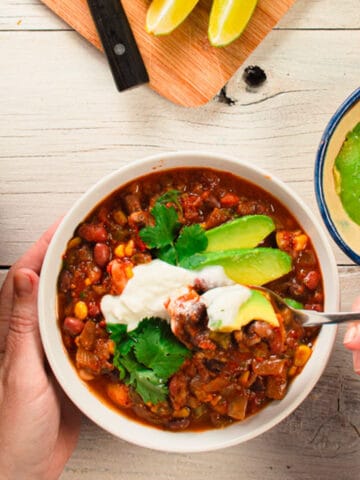 one bowl of chili garnished with avocado, sour cream, and cilantro. hands holding bowl and spoon.