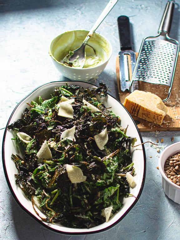 kale caesar salad on white oval platter with parmesan cheese, sunflower seeds and empty dressing bowl on the side