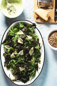kale caesar salad on white oval platter with parmesan cheese, sunflower seeds and empty dressing bowl on the side