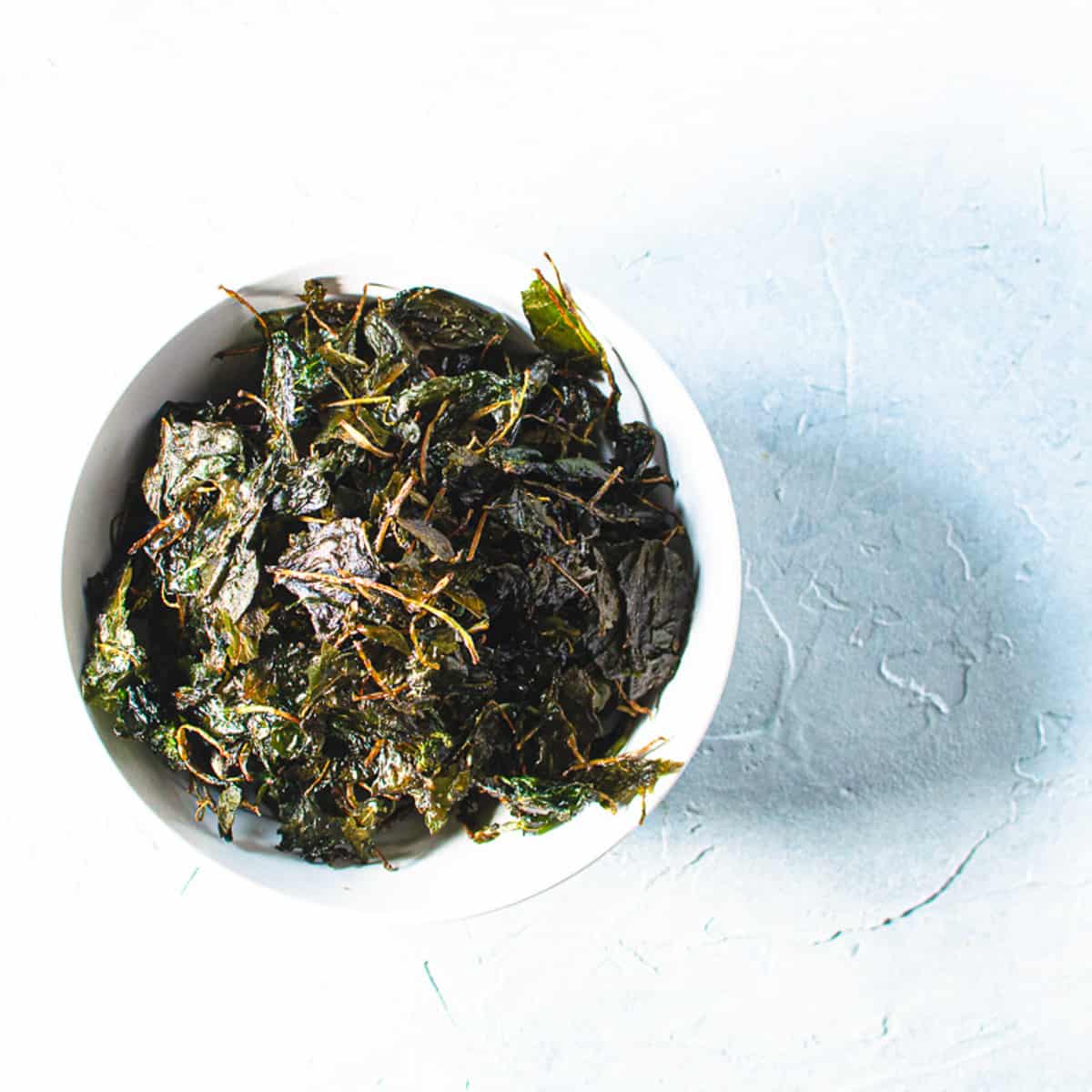 kale crisps in white bowl on light blue background.