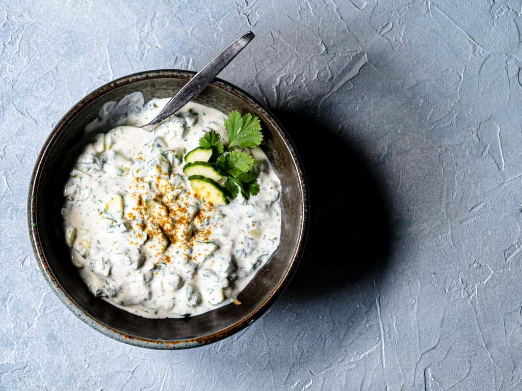 bowl of cucumber raita on light blue background garnished with cucumber slices and cilantro