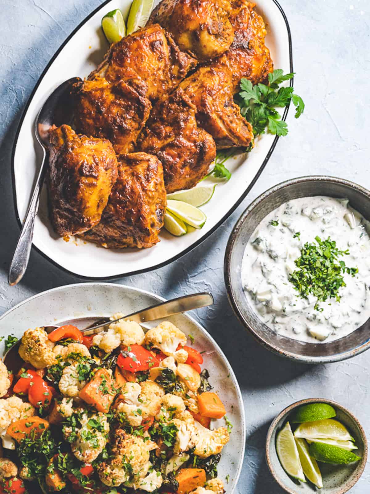 family style platters of curried chicken thighs and roasted vegetables. A dish of cucumber raita and fresh limes on the side. 