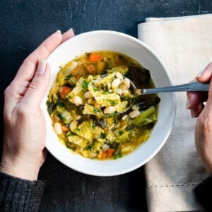 greens and beans soup in a white bowl with a hand cupping one side and a spoon on the other.