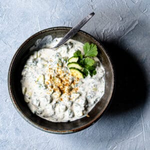 one recipe of raita in a black ceramic bowl garnished with fresh cucumber slices, cilantro and paprika.