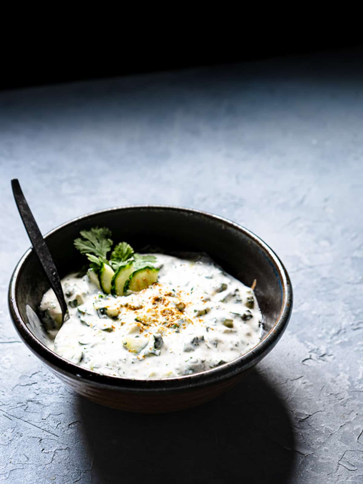 raita in a dark ceramic bowl with a serving spoon. Dish is garnished with cucumber slices and fresh cilantro.