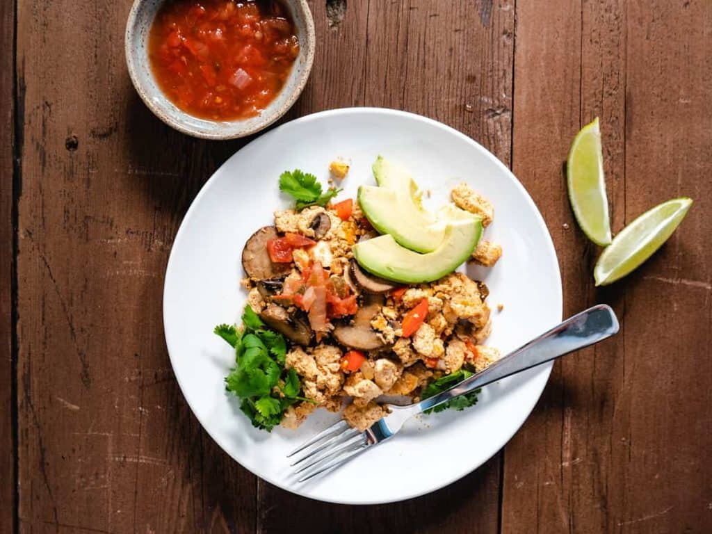 tofu scramble on white plate with avocado slices, lime wedges and a small dish of salsa on the side