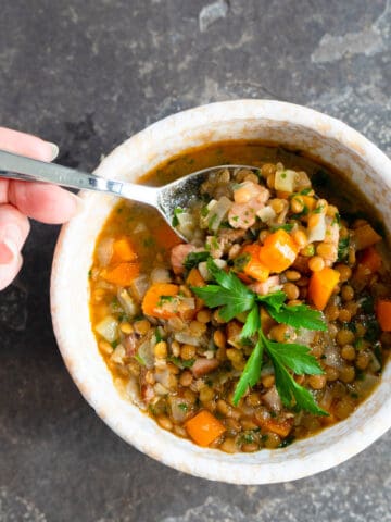 one bowl of lentil soup with a spoon full of soup being held above the bowl.
