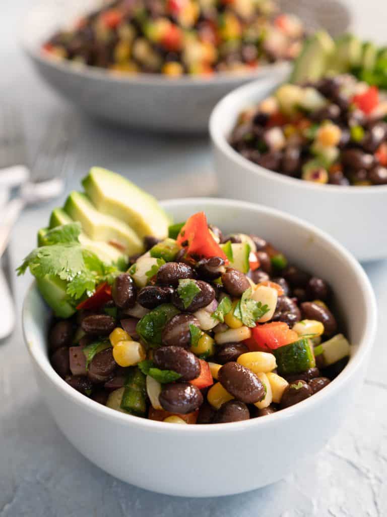 Close up of a white bowl of tex-mex black bean salad with 2 bowls in the background. Garnished with avocado slices