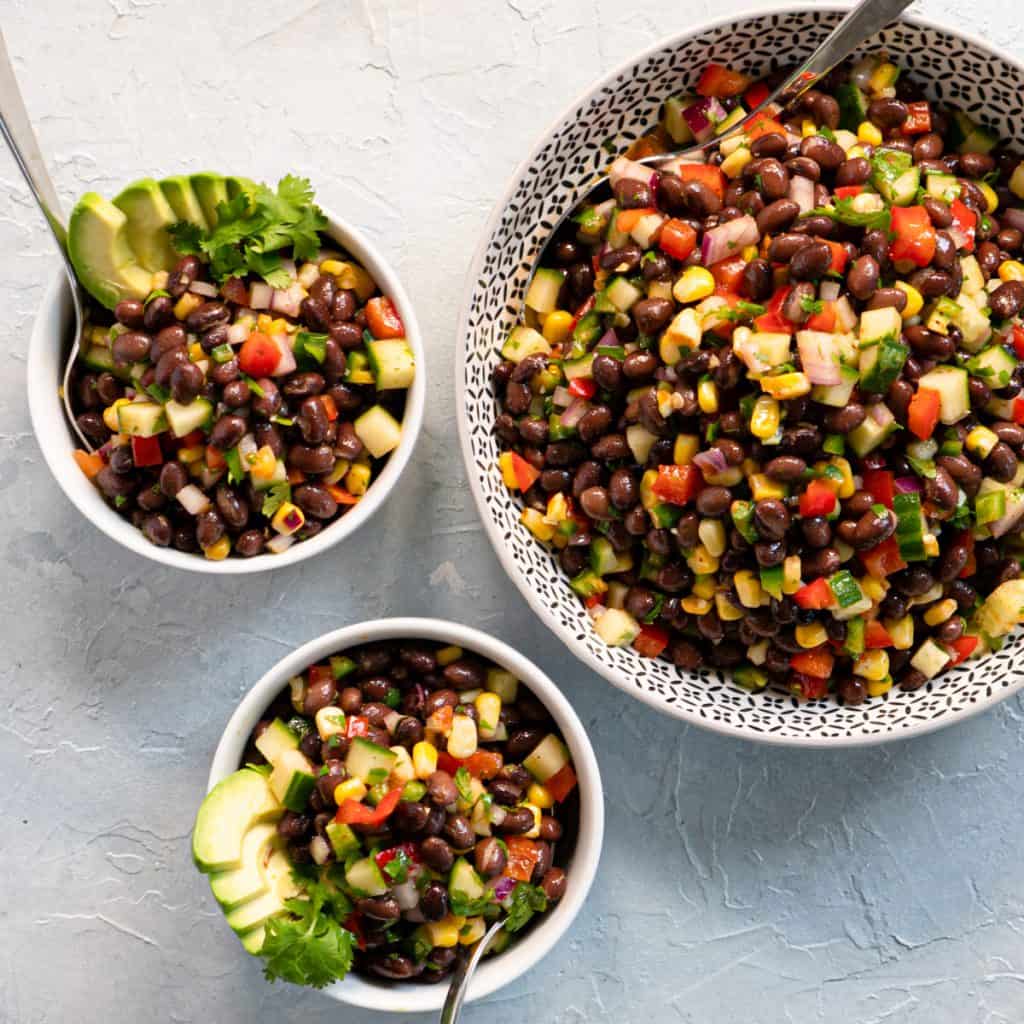 Tex-mex black bean salad in serving bowl with two individual bowls. Garnished with sliced avocado