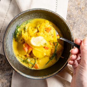 Fish stew with fennel and saffron cream in dark ceramic bowl. A hand holding spoonful of stew