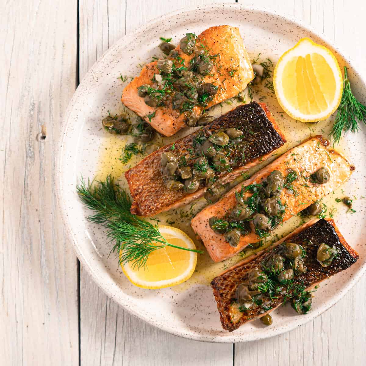 four fillets of Pan seared salmon with fried caper vinaigrette on a white ceramic plate, garnished with fresh dill and lemon.