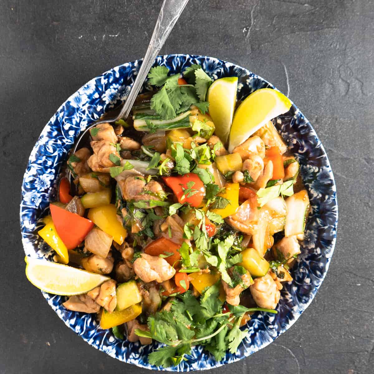 Thai style chicken stir fry in blue and white bowl with spoon. Garnished with fresh lime and cilantro