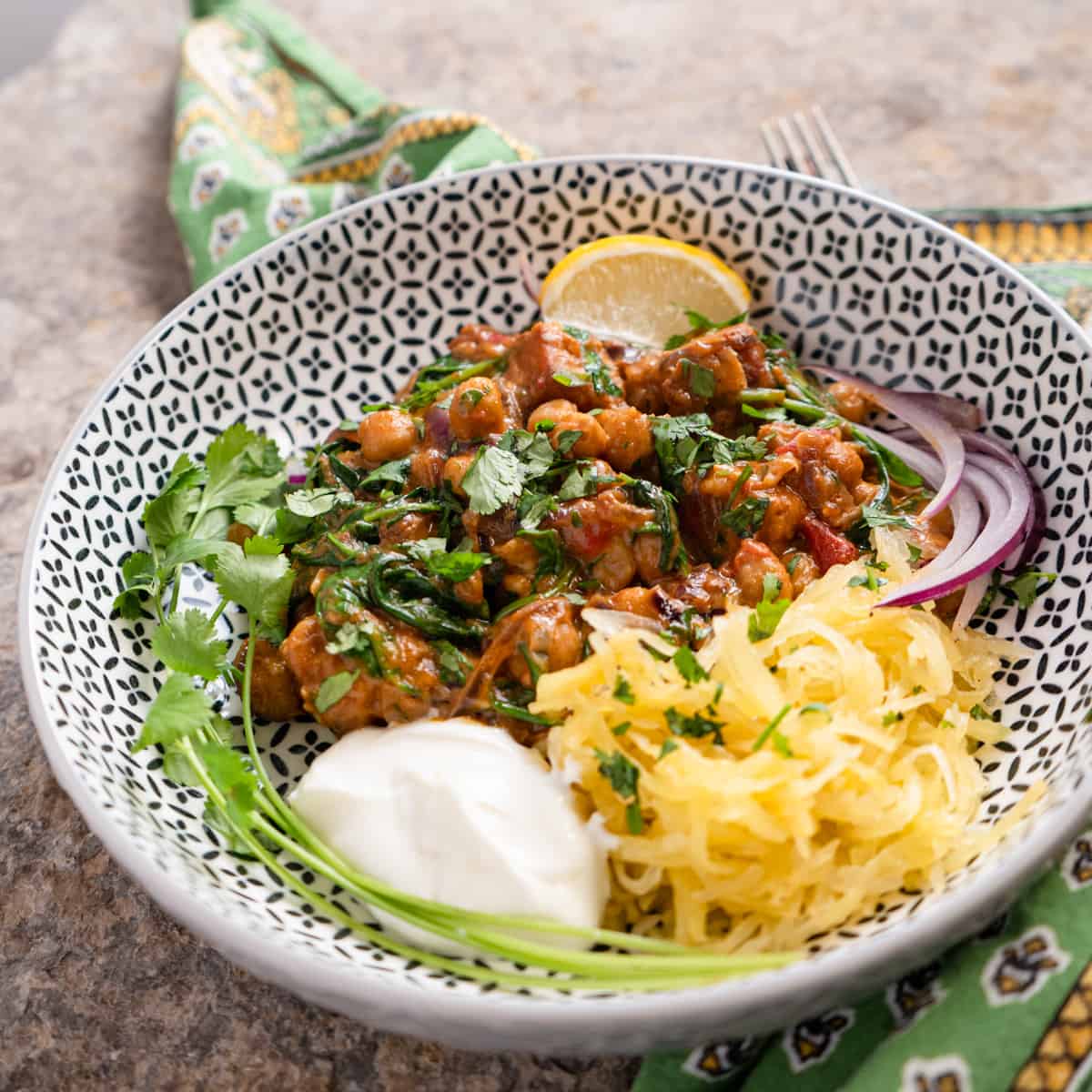 One serving of tempeh curry in a bowl with spaghetti squash, yogurt, red onions and fresh lemon on the side. 