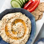 Prepared hummus in a blue dish with sliced cucumber, red pepper and crackers on the side.