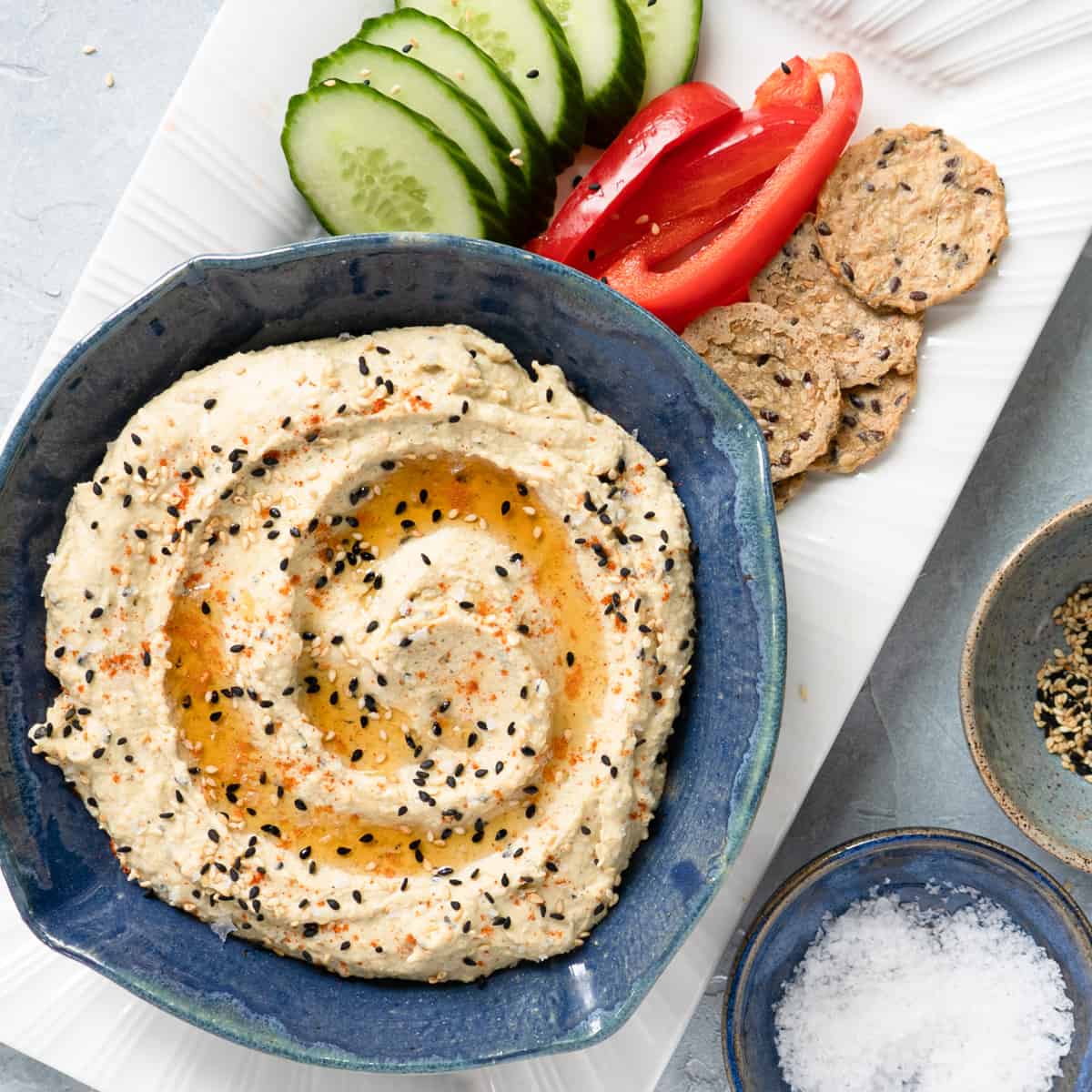 Prepared hummus in a blue dish with sliced cucumber, red pepper and crackers on the side.