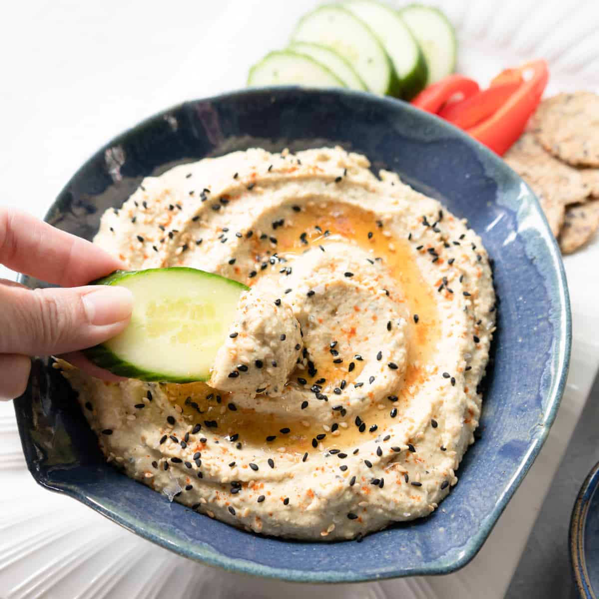 Prepared hummus in a blue dish with a hand dipping a slice of cucumber into it. 