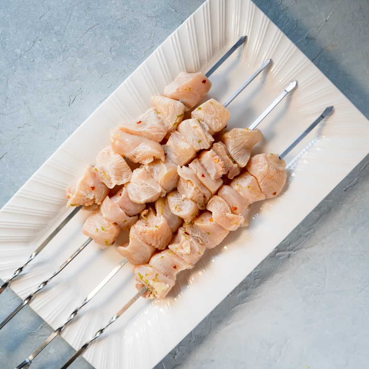 4 chicken kabobs on a white plate before grilling.
