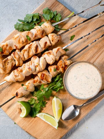 Cooked chicken kabobs on a board with dipping sauce, lime wedges, and fresh cilantro.