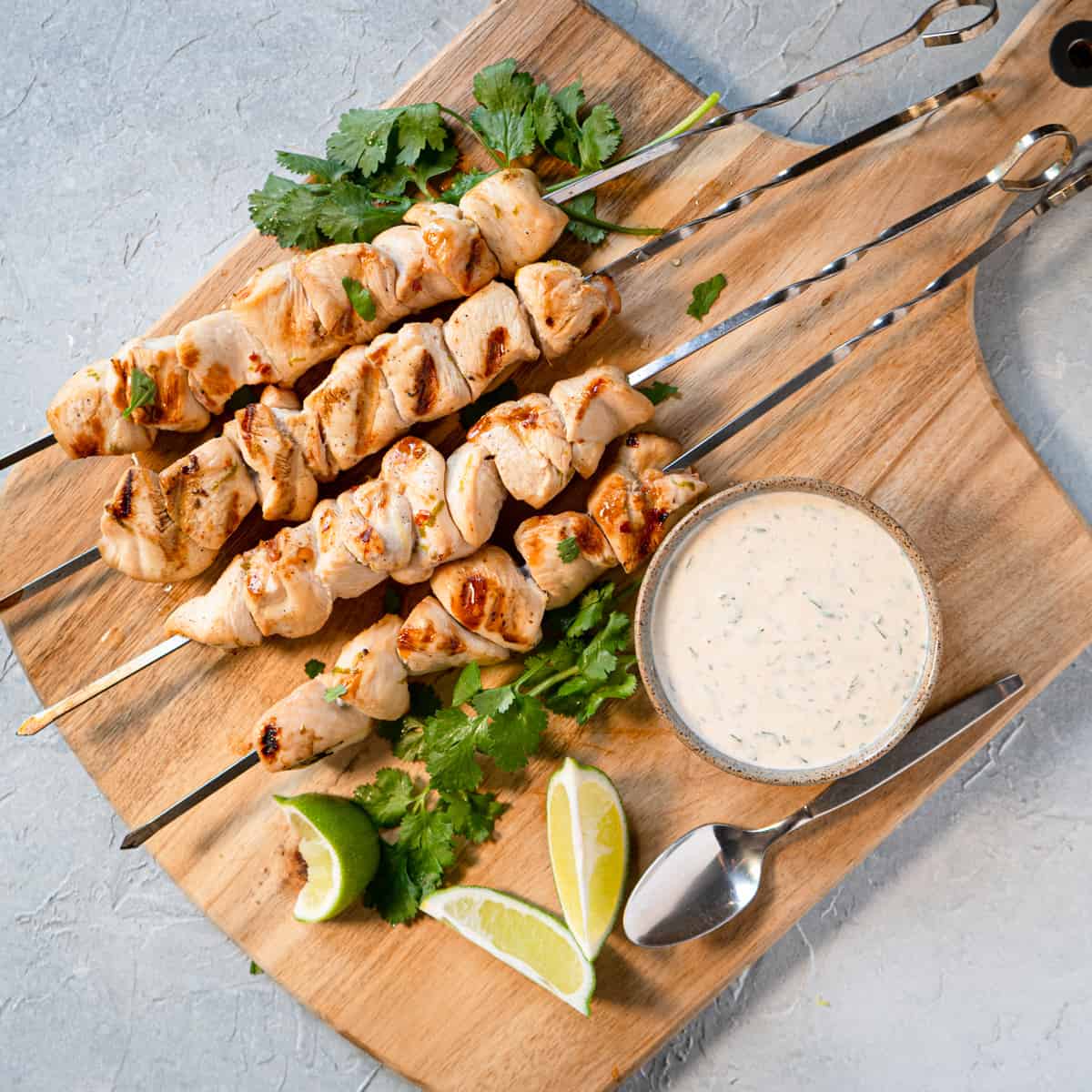 Cooked chicken kabobs on a board with dipping sauce, lime wedges, and fresh cilantro.