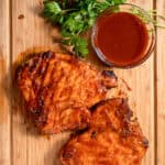 2 grilled pork chops on a wood board with a side of bbq sauce and parsley garnish.