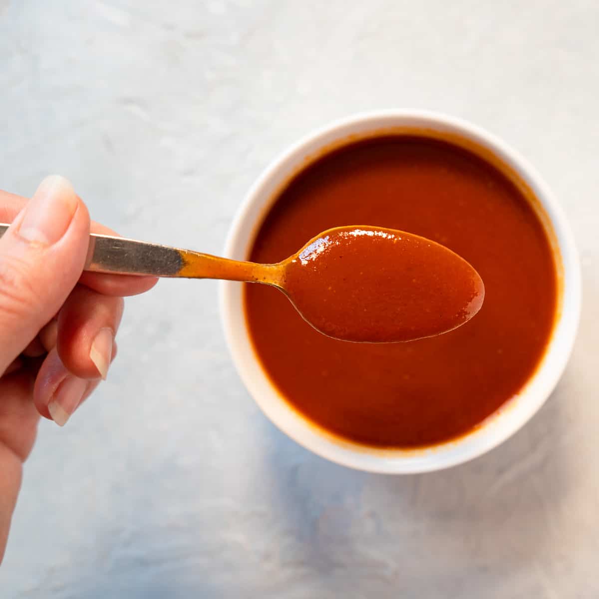Prepared bbq sauce in a white bowl with a hand holding a spoonful to show how smooth it is.