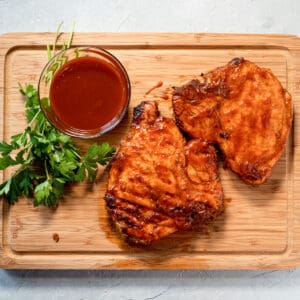2 grilled pork chops on a wood board with a side of bbq sauce and parsley garnish.