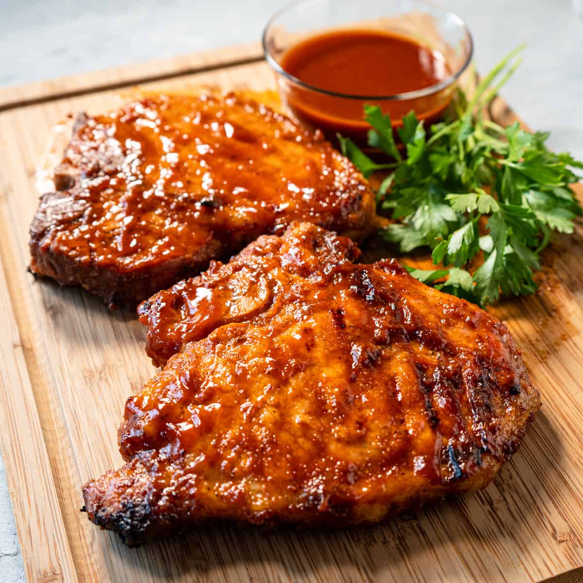 Grilled pork chops on a cutting board with a side of sauce and parsley garnish. 