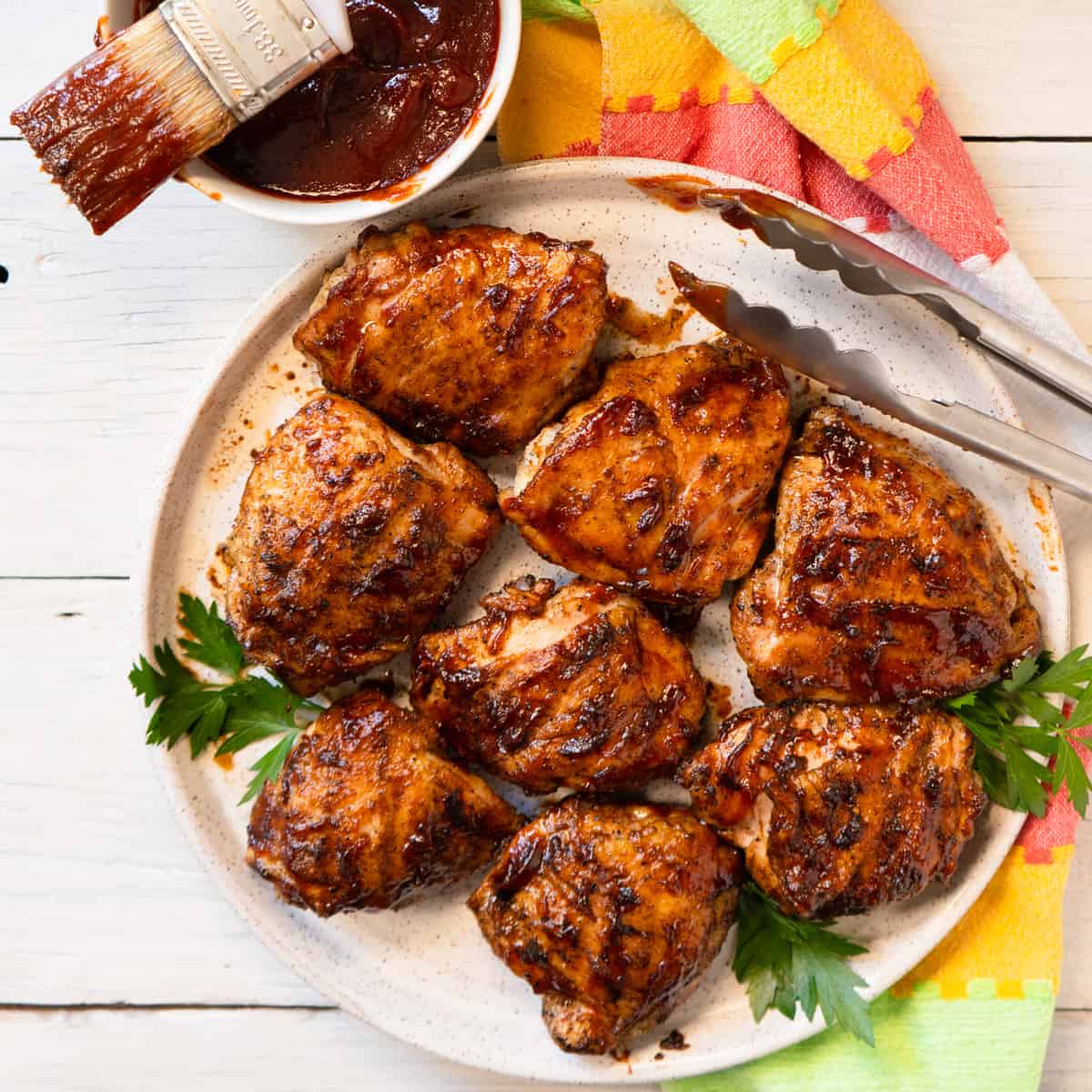 4 bbq chicken thighs on a white plate with extra sauce on the side.