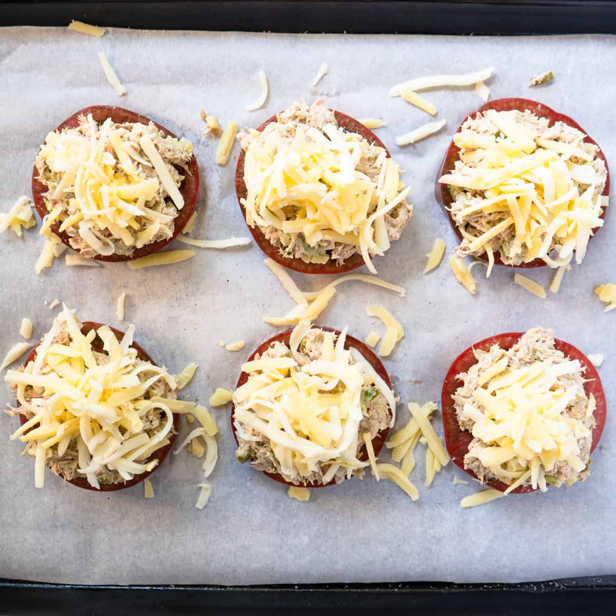 six slices of tomato topped with tuna salad and cheese on a baking tray.