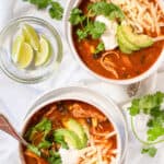 two bowls of enchilada soup topped with shredded cheese, avocado, sour cream, and fresh cilantro.