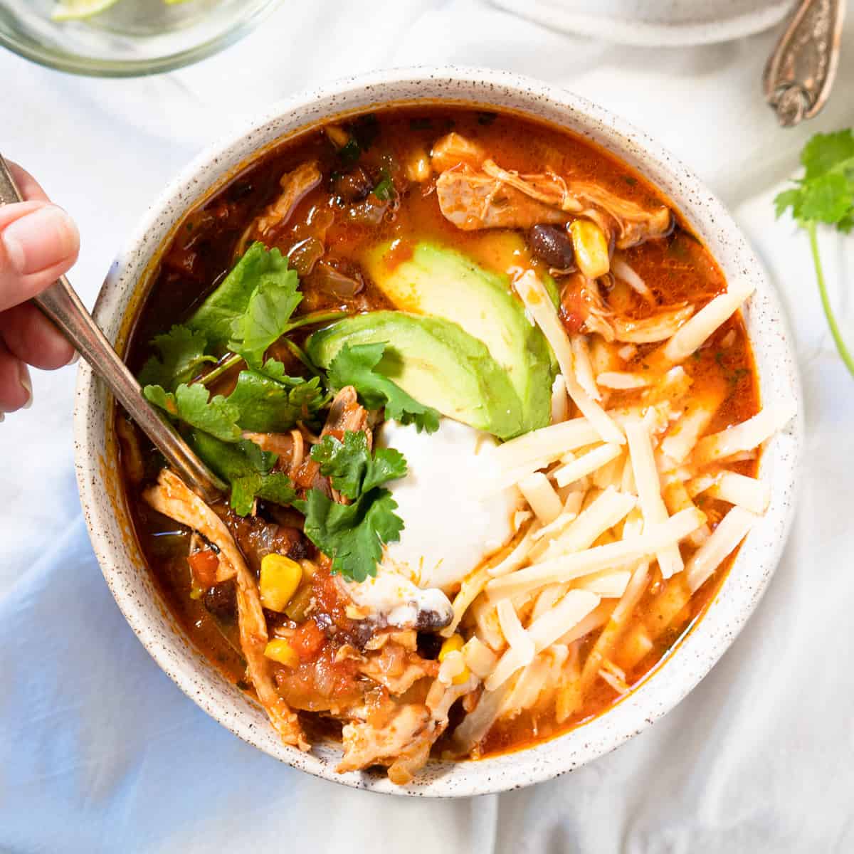 Single serving of chicken enchilada soup topped with cheese, avocado, sour cream. Hand holding a spoonful of soup. 