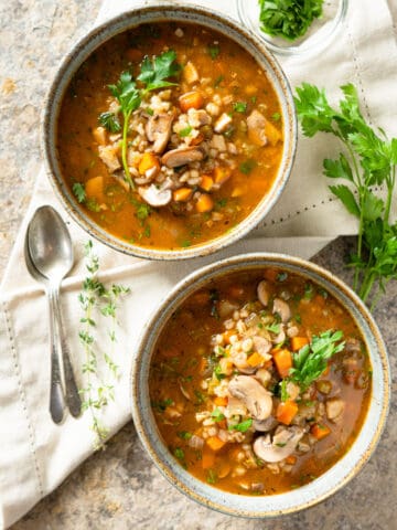 2 bowls of mushroom barley soup with spoons on the side garnished with fresh parsley.