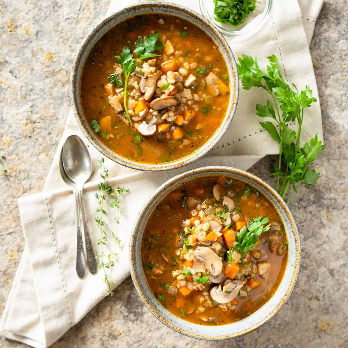 2 bowls of mushroom barley soup with spoons on the side garnished with fresh parsley.