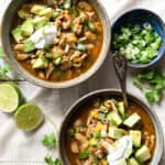 2 bowls of green chicken chili garnished with avocado, sour cream, and green onions.