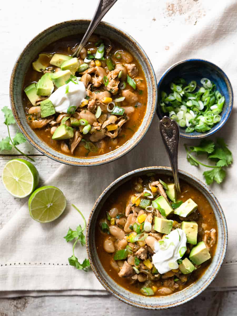 2 bowls of green chicken chili garnished with avocado, sour cream, and green onions.