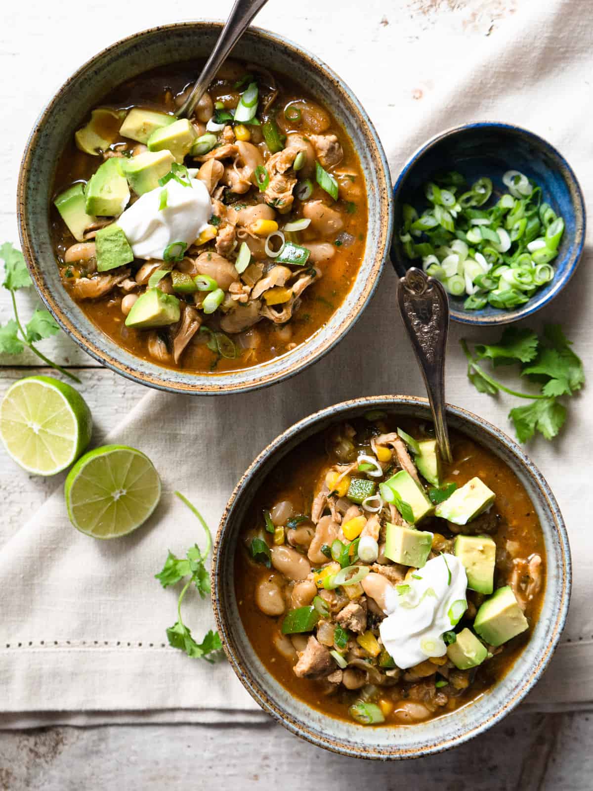 2 bowls of green chicken chili garnished with avocado, sour cream, and green onions.