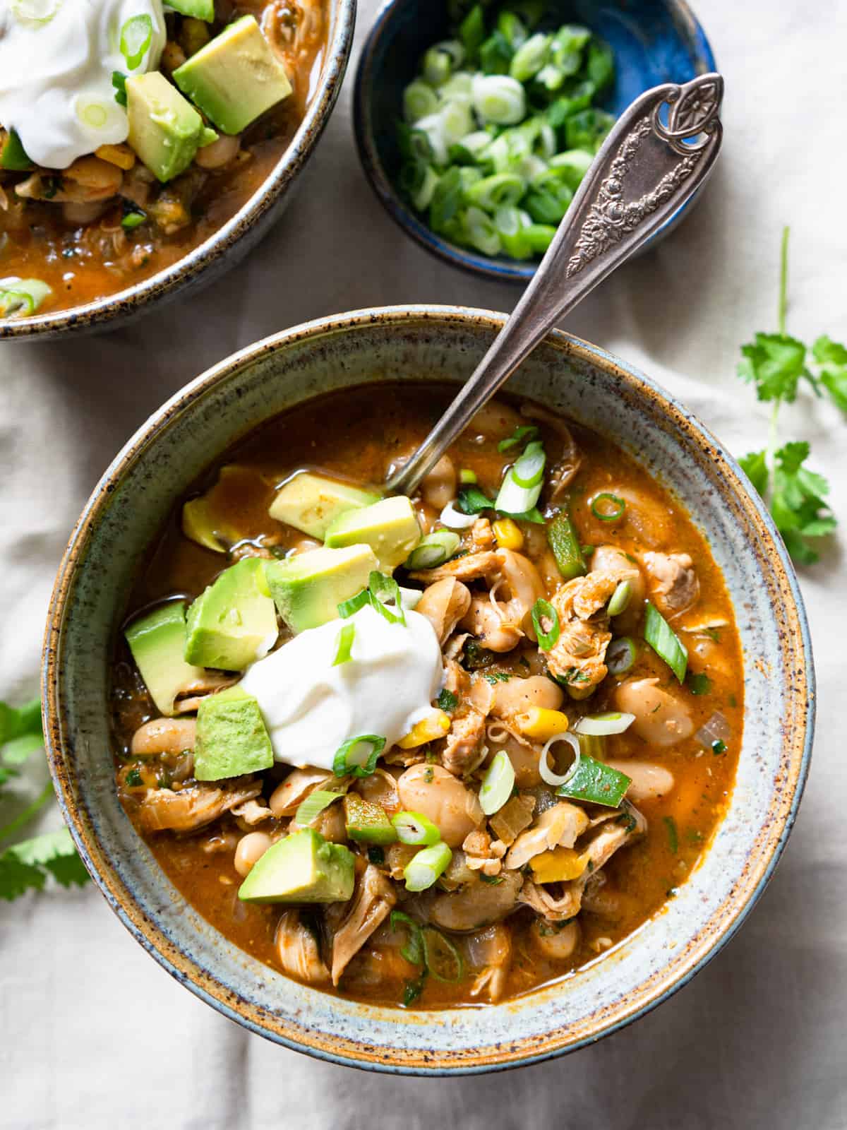 one serving of chili in a ceramic bowl with a spoon and garnished with avocado, sliced green onions and sour cream. 