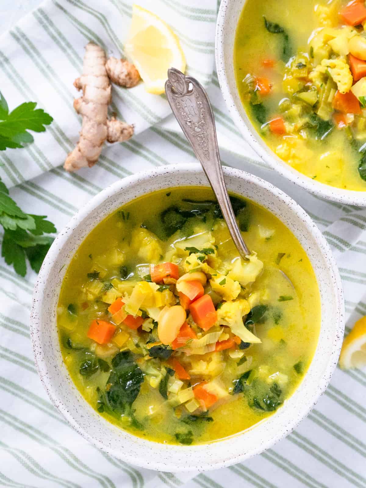 one white bowl of detox soup with a second on the side. Fresh turmeric root and lemon wedge on the other side.