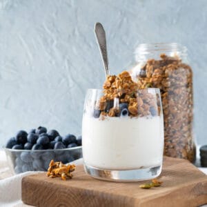 A glass of plain yogurt topped with granola and blueberries. A jar of granola and a dish of blueberries are on the side.